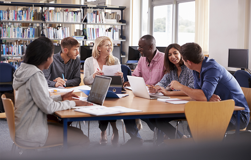 Adults with happy engaged expressions having a discussion sitting around a desk with laptops and papers in a library setting
