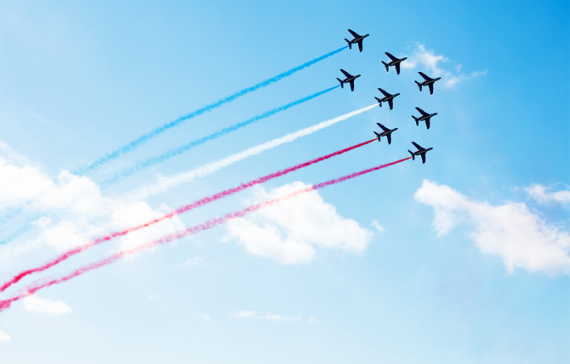 Aerial display by fighter jets: eight small jets fly in tight formation leaving coloured jet trails behind them, in a clear sky with a few small clouds. A feeling of energy, dynamism, optimism, agility, coordination, team work, and strength.
